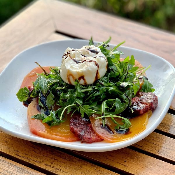 A burrata salad with arugula, sliced tomatoes, and balsamic glaze on a white plate.
